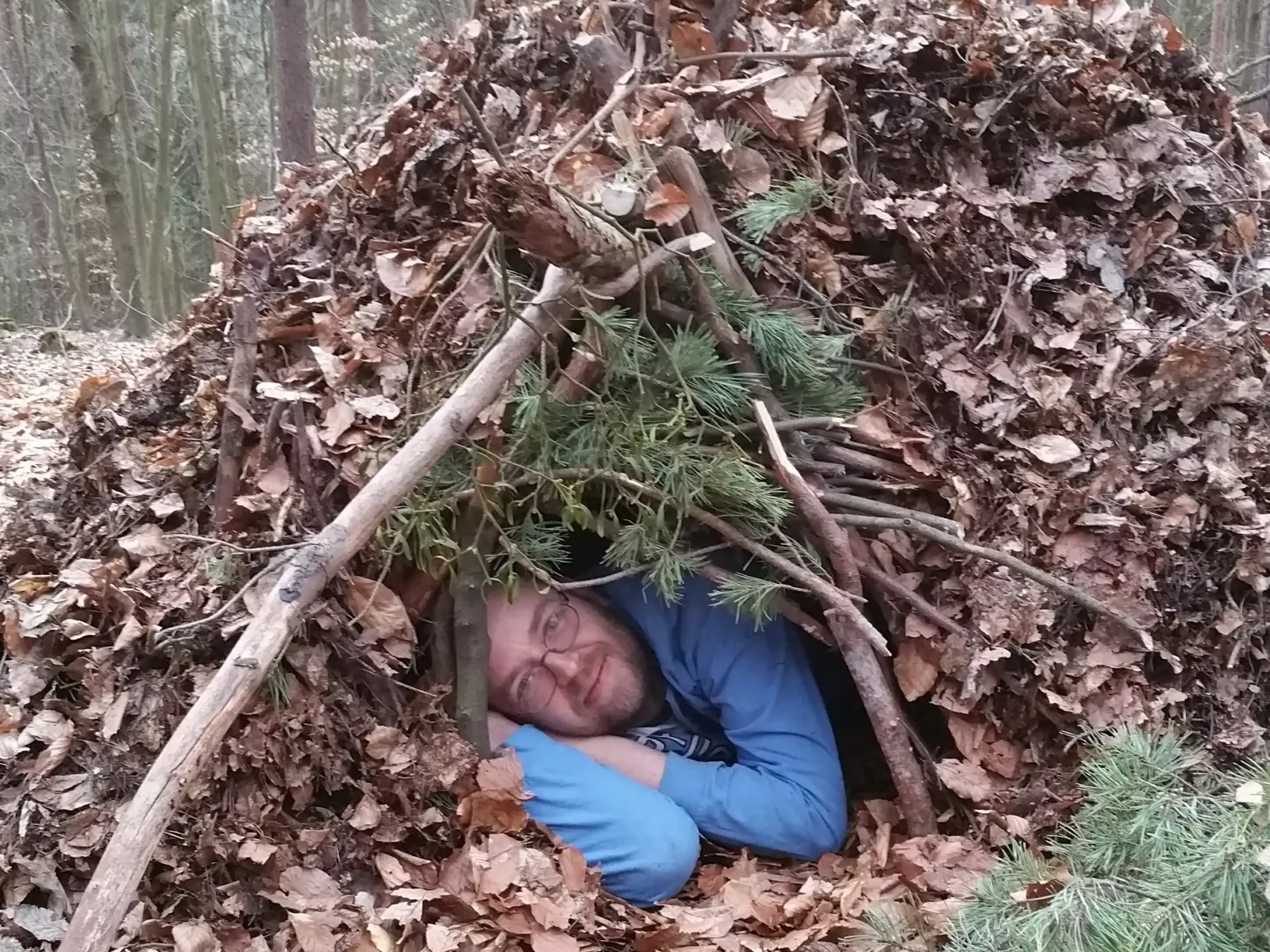 Ein Mann liegt in einer selbstgebauten Laubhütte im Wald beim Survivaltag.