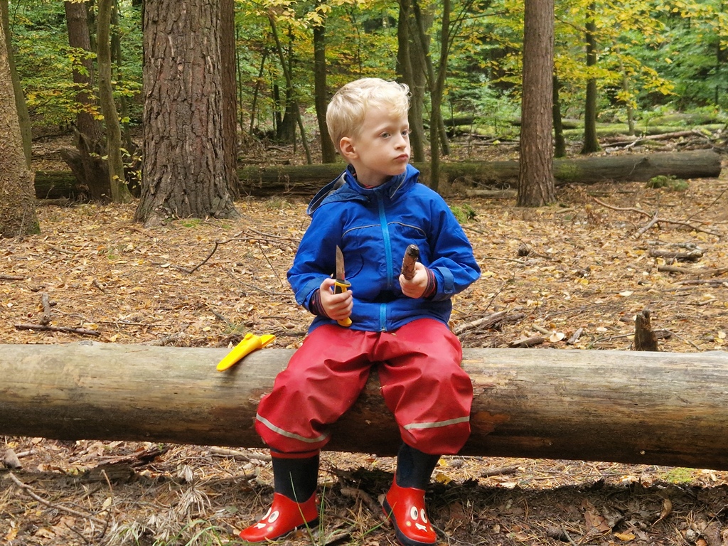 Eine Junge sitzt auf einem Baumstamm im Wald und schnitzt.