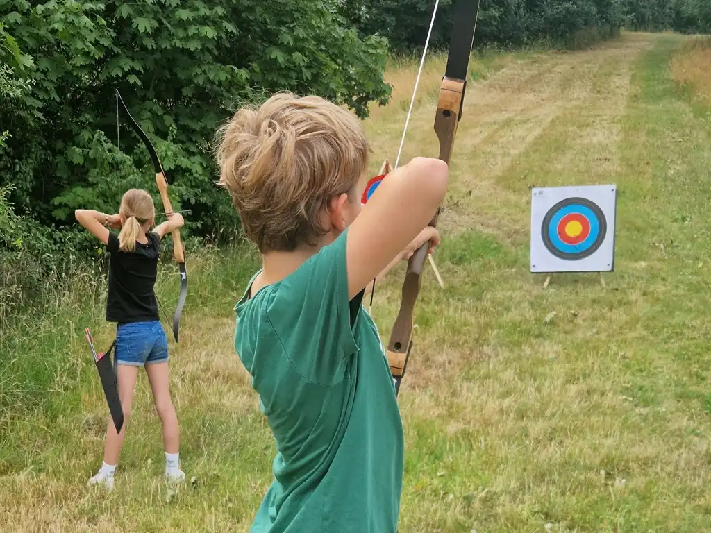 Zwei Kinder schießen mit dem Bogen auf eine Zielscheibe beim wilden Kindergeburstag.